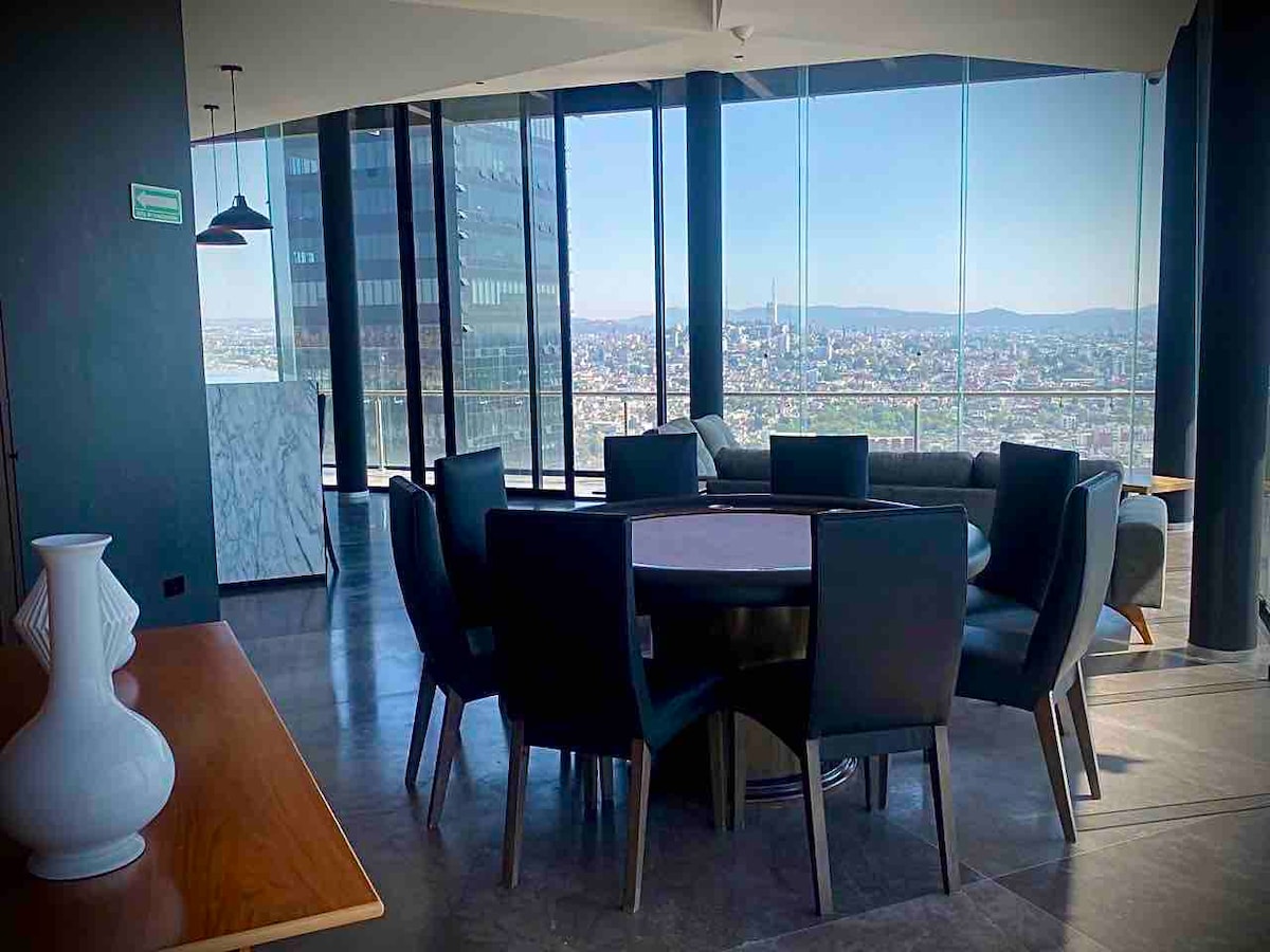 A bright dining area is visible, featuring a round table surrounded by black chairs. Large windows extend from the floor to the ceiling, providing expansive views of the city and mountains in the distance. A marble accent wall complements the modern design.