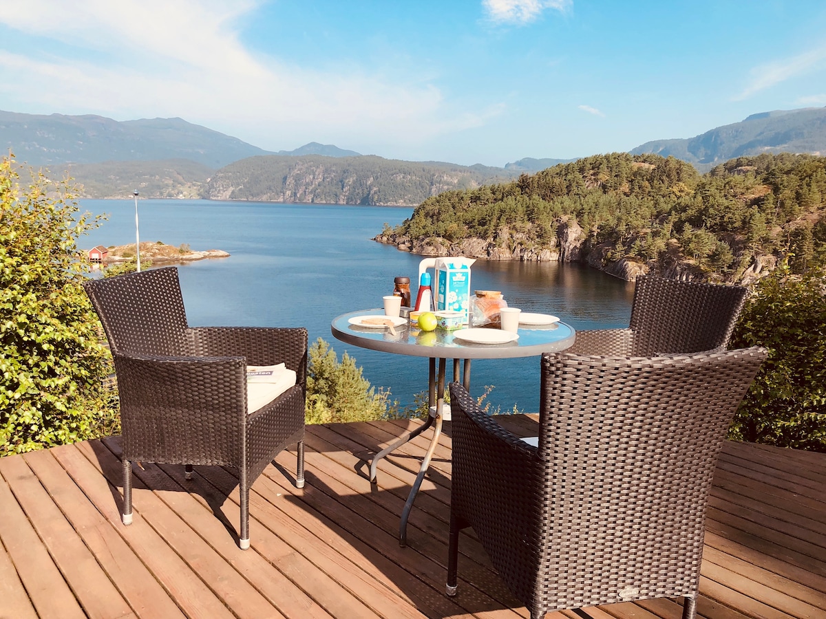 A scenic outdoor dining area features two wicker chairs and a round table, set against a backdrop of a serene fjord. The table is adorned with breakfast items, and expansive views of the water and hills are visible in the distance.