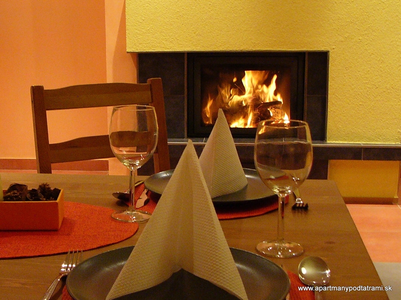A dining table is set with plates and glasses, accompanied by neatly folded napkins. A warm glow emanates from the fireplace in the background, against a textured yellow wall, creating a cozy environment suitable for relaxed dining.