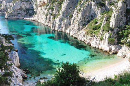 A calm cove is framed by steep, rocky cliffs, showcasing clear turquoise waters. A sandy beach is visible at the shoreline, bordered by lush green vegetation. The tranquil scene captures the natural beauty of the coastline, inviting peaceful relaxation and exploration.