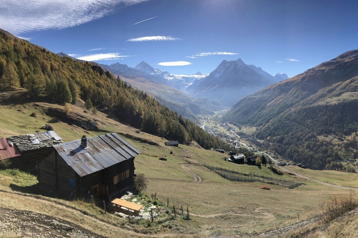 A rustic mountain chalet is positioned on a hillside, featuring weathered wooden exterior and a spacious outdoor table. Surrounded by rolling landscapes, it overlooks a vast valley framed by majestic mountains under a clear blue sky, showcasing the natural beauty of the Alps.