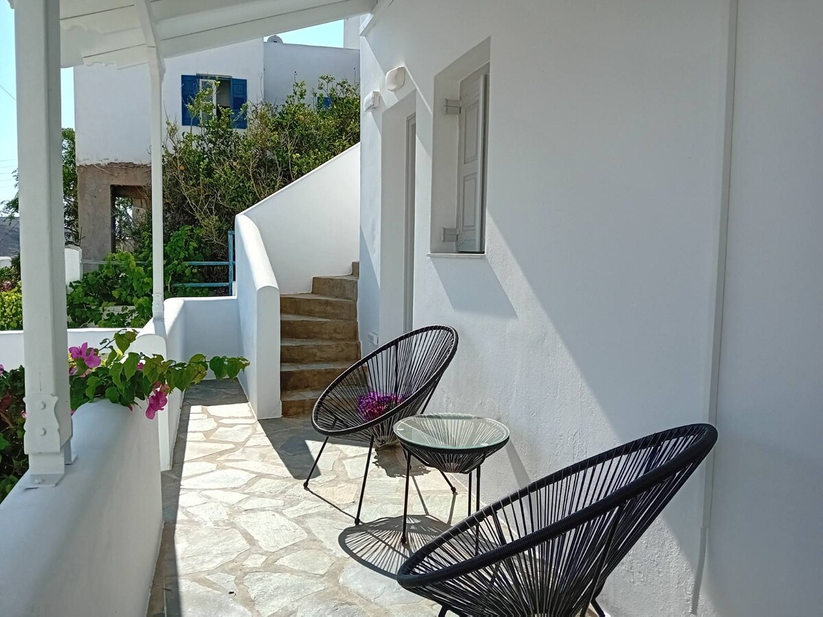 A sunlit terrace features two black woven chairs positioned around a small glass table. Vibrant green plants and flowers line the space, while stairs lead to the entryway of the building, which is framed by soft white walls.