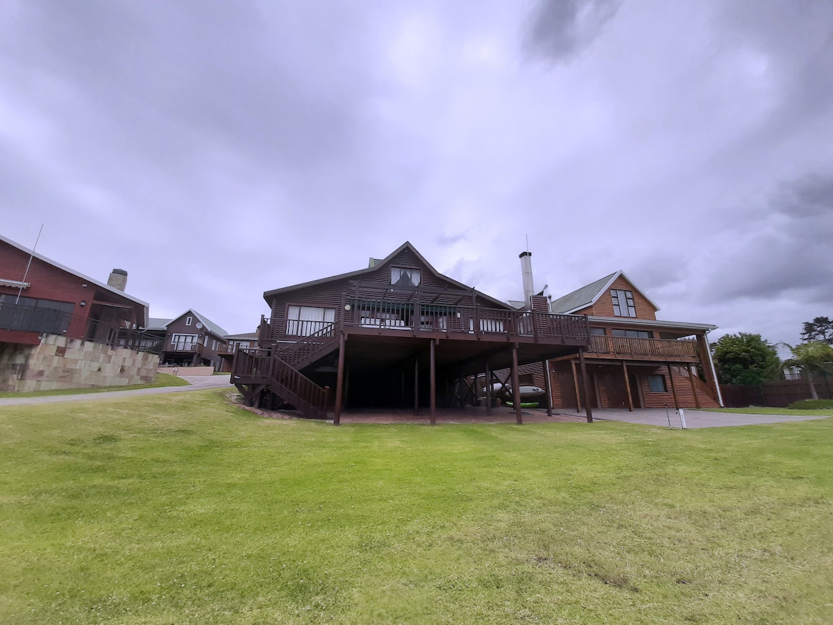 A wooden log home is set on an expansive lawn, showcasing a broad deck that extends out toward the surroundings. The two-story structure has multiple gables and windows, reflecting the rustic charm of the lodge. Nearby buildings and pathways are also visible.
