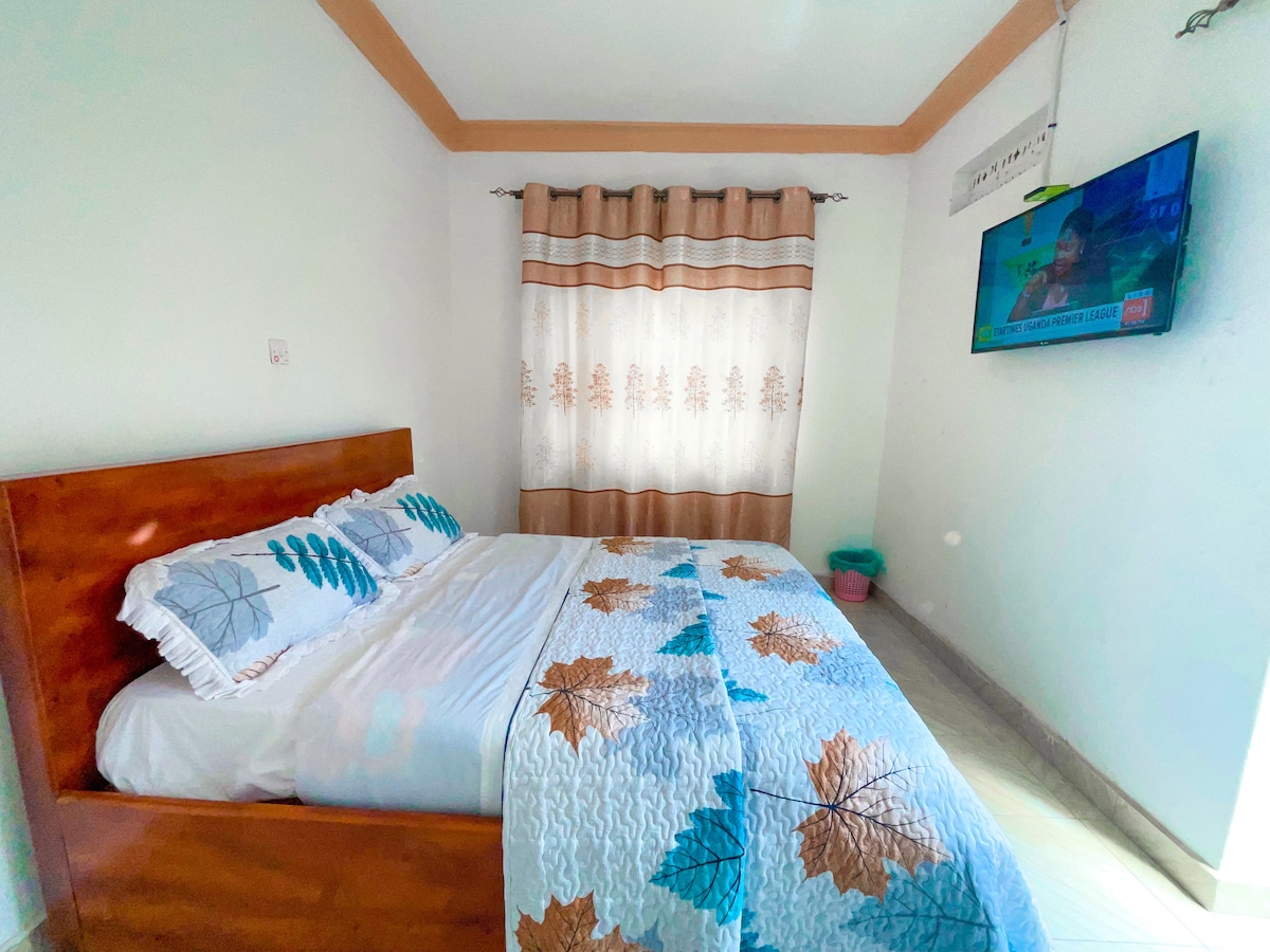 A comfortable bedroom features a wooden bed dressed with a patterned blanket. A large window with light-colored curtains allows natural light to filter in. A television mounted on the wall displays a news channel, contributing to the room’s functionality.