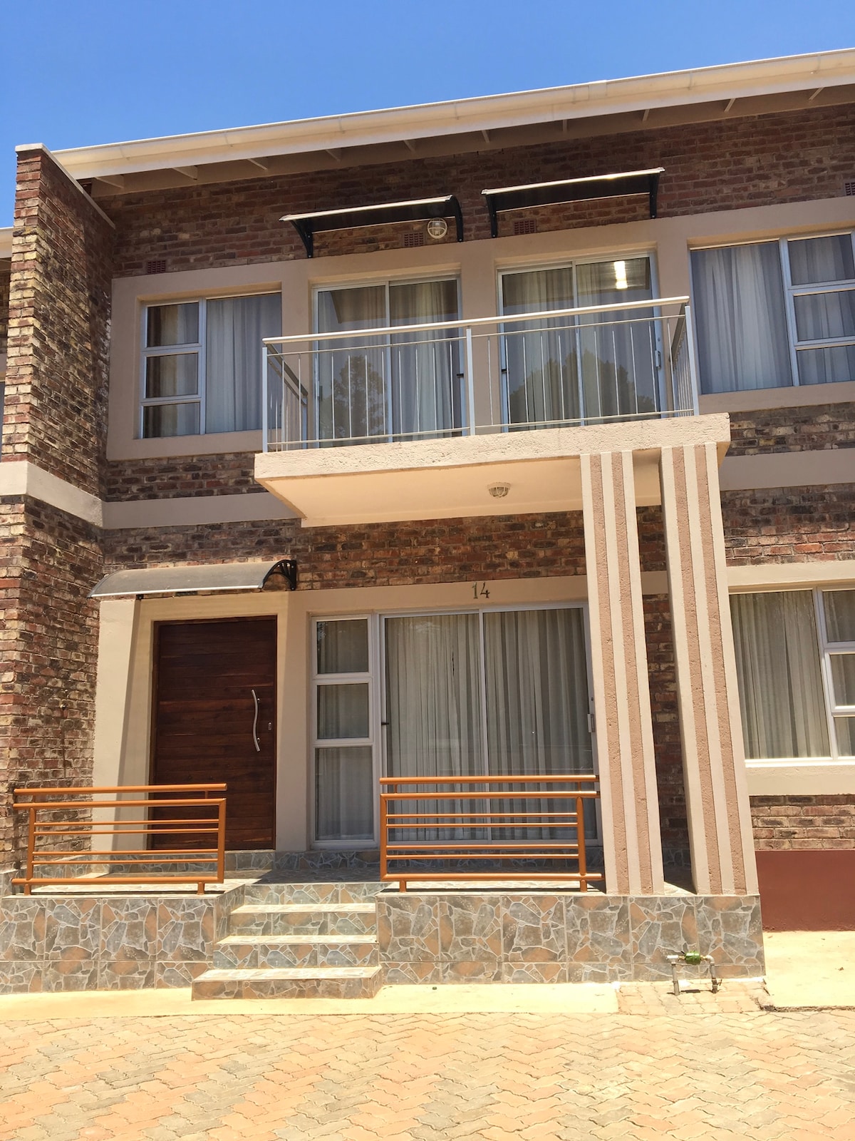 The apartment entrance features a stone pathway leading to a wooden front door, with a small balcony extending over the entrance. Large windows with sheer curtains provide natural light, and the brick façade complements the modern detailing of the structure.