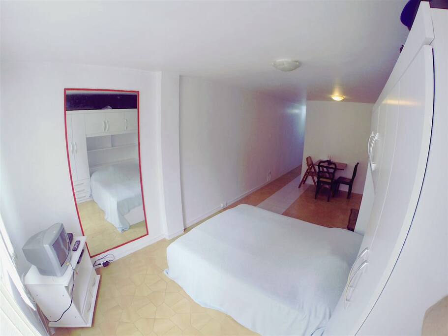 Cama de casal, corredor e mesa ao fundo.