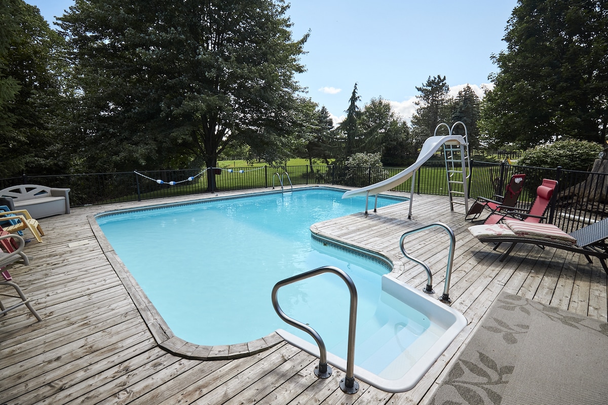 A heated swimming pool is surrounded by wooden decking, featuring a slide and handrails for access. Colorful lounge chairs are positioned nearby, and a hammock is visible in the background, with lush greenery creating a tranquil outdoor setting.