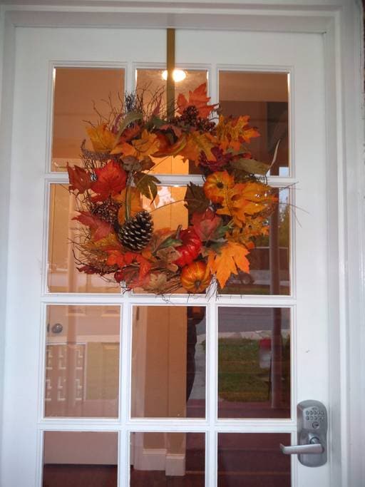 A decorative autumn wreath composed of vibrant orange and yellow leaves, pine cones, and small gourds is displayed on a glass-panelled door. Light from inside the building illuminates the entryway, enhancing the welcoming appearance.