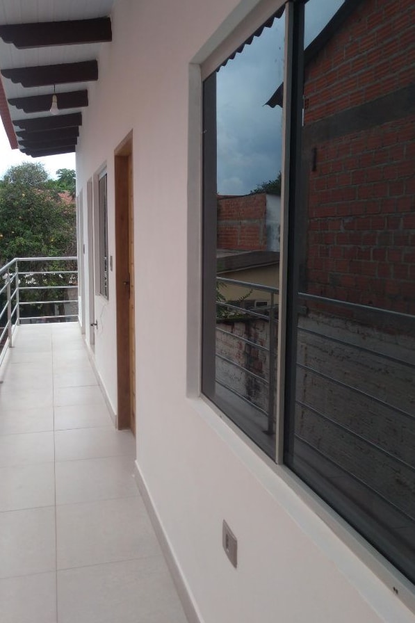 An outdoor balcony space is presented, featuring smooth tile flooring. Large windows reflect the surrounding greenery, while a door leads into the interior space. A railing offers safety and maintains openness, with a view of a brick building in the background.