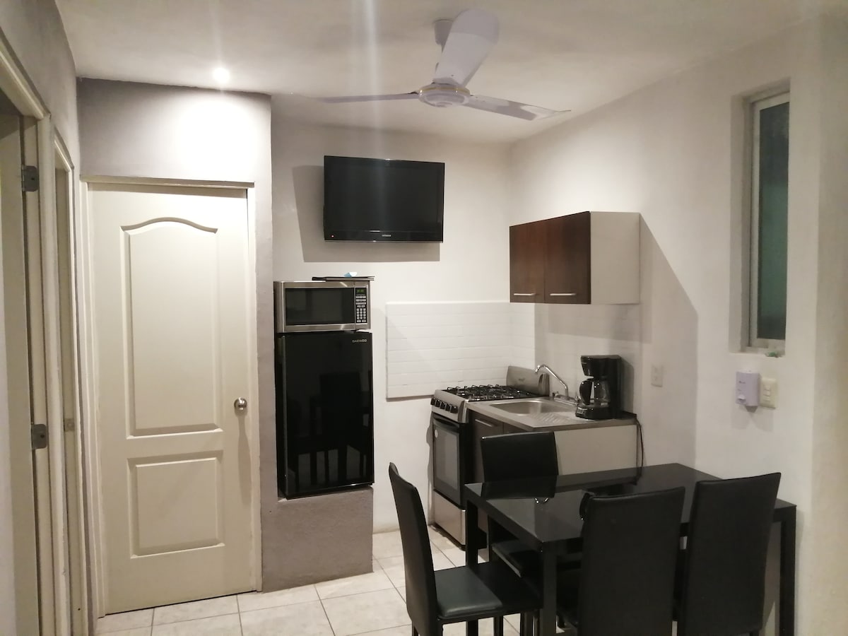 A kitchen area is visible, featuring a black refrigerator and a stove with an oven. A dining table with six chairs is positioned nearby. A flat-screen television is mounted on the wall, and a window allows natural light to enter.