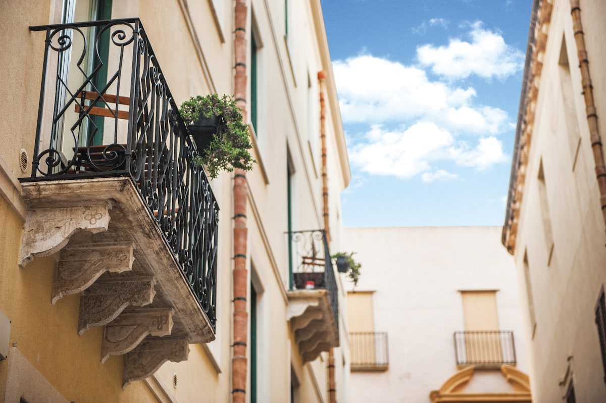 A wrought-iron balcony extends from a pale yellow building, adorned with potted plants. The sky above is scattered with clouds, framing the serene urban scene. Additional balconies can be seen on nearby buildings, contributing to the architectural charm of the area.