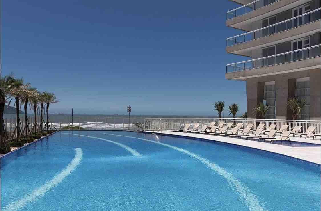 An expansive outdoor pool reflects the clear blue sky, bordered by lounge chairs for relaxation. Lush palm trees line the pool area, enhancing the coastal environment. The ocean is visible in the background, further emphasizing a serene setting.