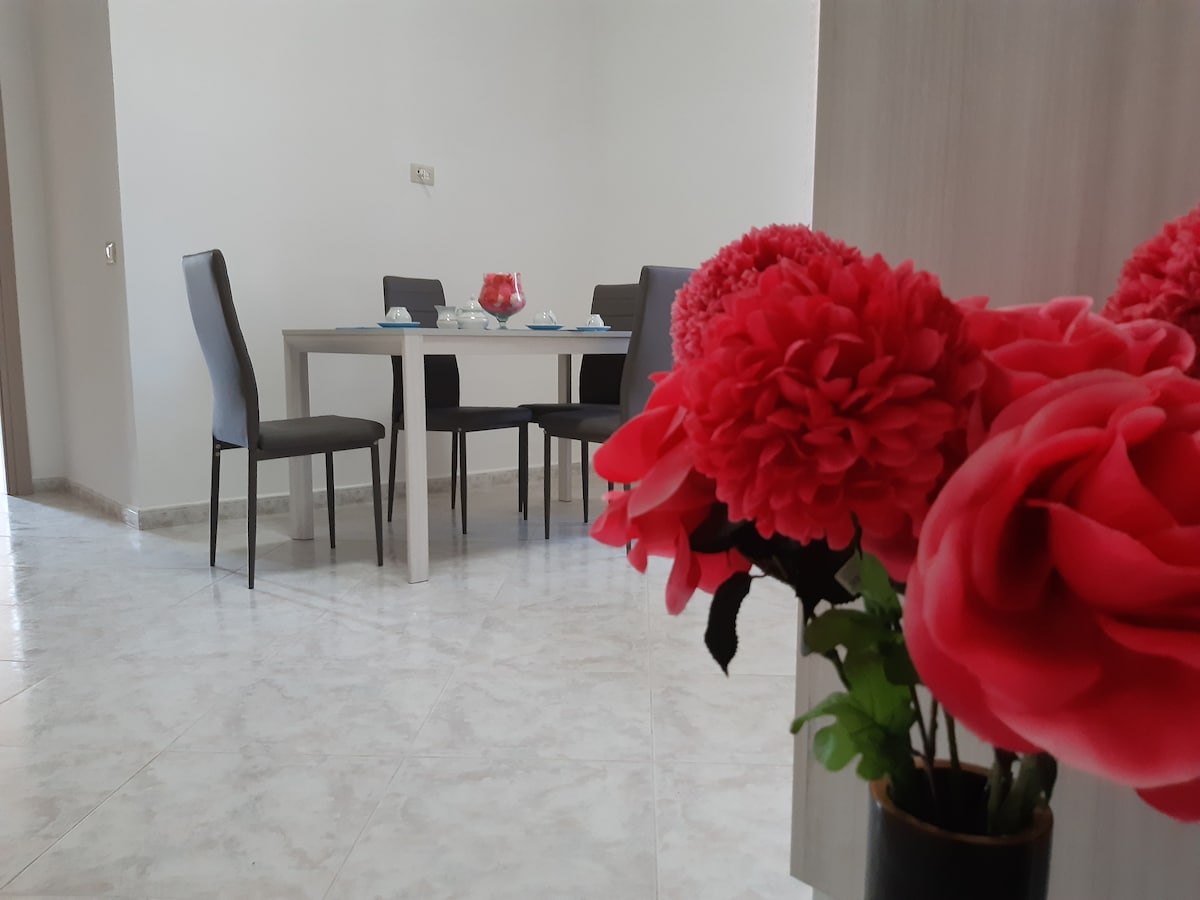 A dining area is visible, featuring a rectangular table set for a meal with six chairs. Bright red flowers in a vase add a splash of color, while the light-colored flooring enhances the spacious feel of the room.