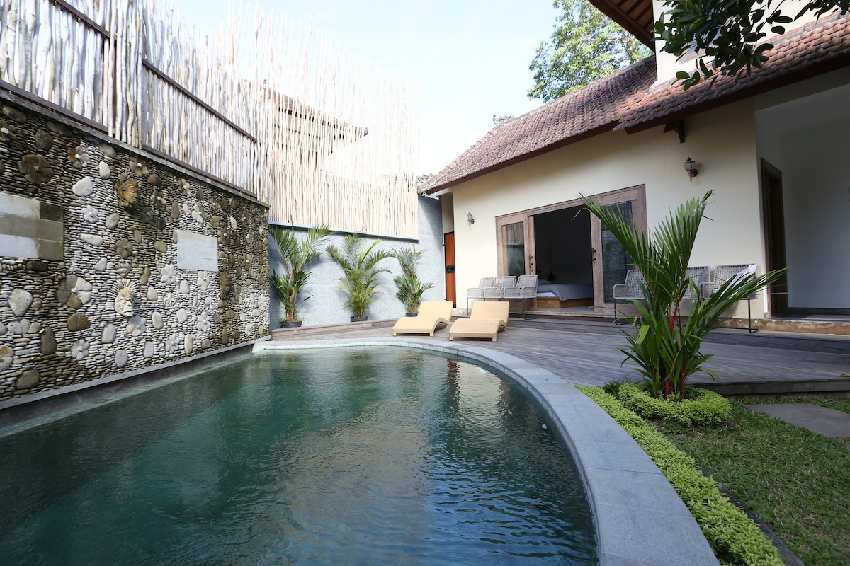 An inviting private garden is highlighted by a reflective swimming pool, surrounded by green landscaping. Sun loungers are positioned near the water, and a modern, two-story villa can be seen in the background, with an outdoor deck seamlessly blending with the natural environment.