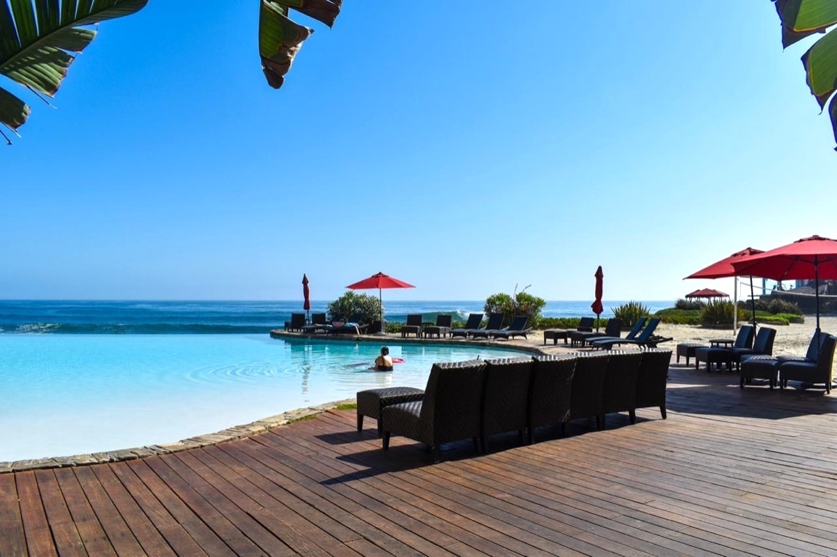 An oceanfront infinity pool is featured, blending seamlessly with the horizon. Comfortable lounge chairs are arranged along the wooden deck, shaded by red umbrellas. Palm leaves are visible in the foreground, framing the serene scene with clear blue skies above and gentle waves in the background.