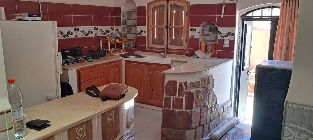 A functional kitchen is presented, featuring wooden cabinetry and a stone countertop. Cooking appliances and utensils are neatly arranged. A window allows natural light, while an entrance leads to an outdoor area. The decor includes warm tones and subtle patterned tiles.