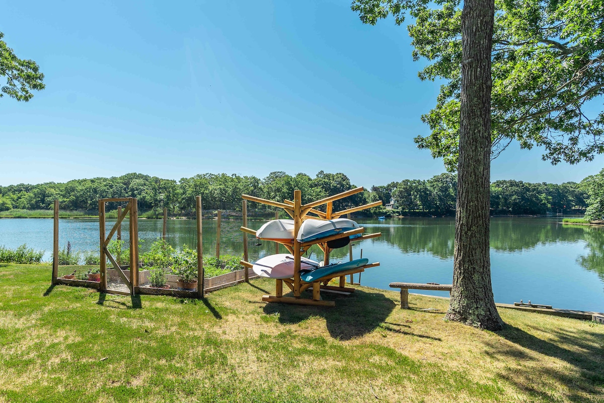 A serene waterfront view of Hashamomuck Pond is showcased, with kayaks and paddle boards arranged on a wooden rack. Lush greenery surrounds the calm water, complemented by a small garden structure nearby. Sunlight reflects off the water, creating a tranquil atmosphere.