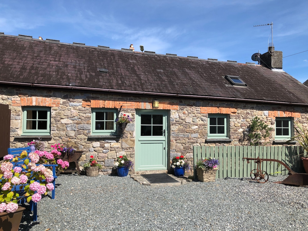 The entrance of a stone cottage is framed by colorful flower pots, showcasing vibrant blooms. The cottage features a pale green door and several windows with dark green frames, set against a backdrop of a slate roof under a bright blue sky.