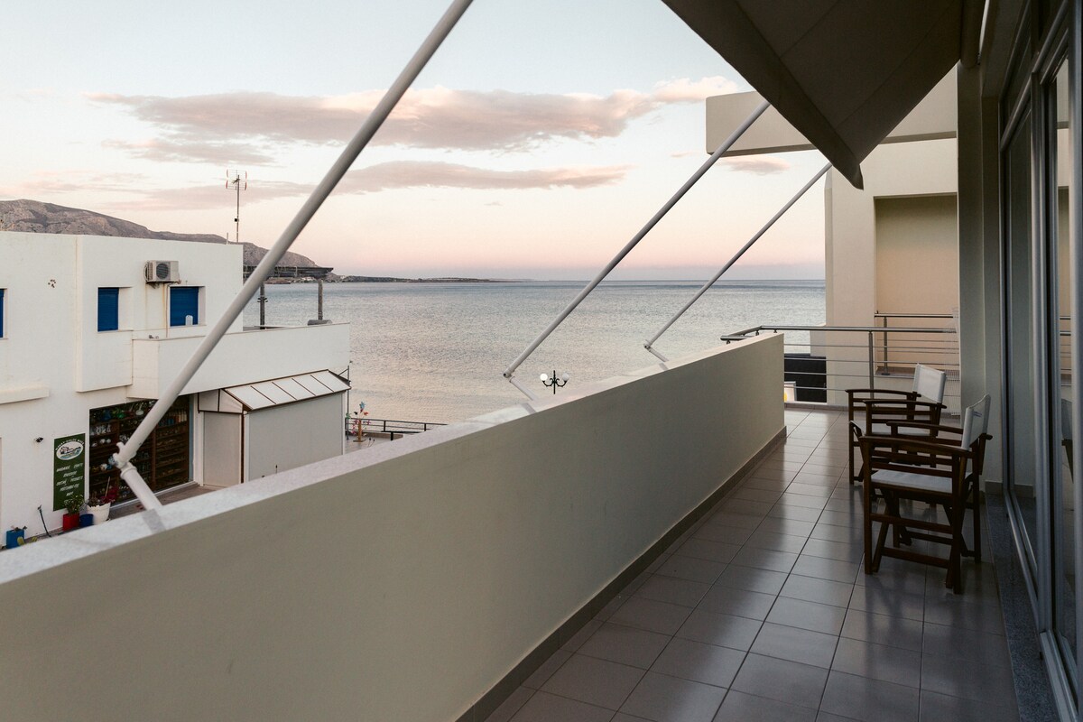 A spacious balcony offers a view of the calm sea at dusk. Two wooden chairs are positioned for relaxation, while the surrounding area features modern architecture and distant hills. The tile flooring adds a clean, contemporary feel to the outdoor space.
