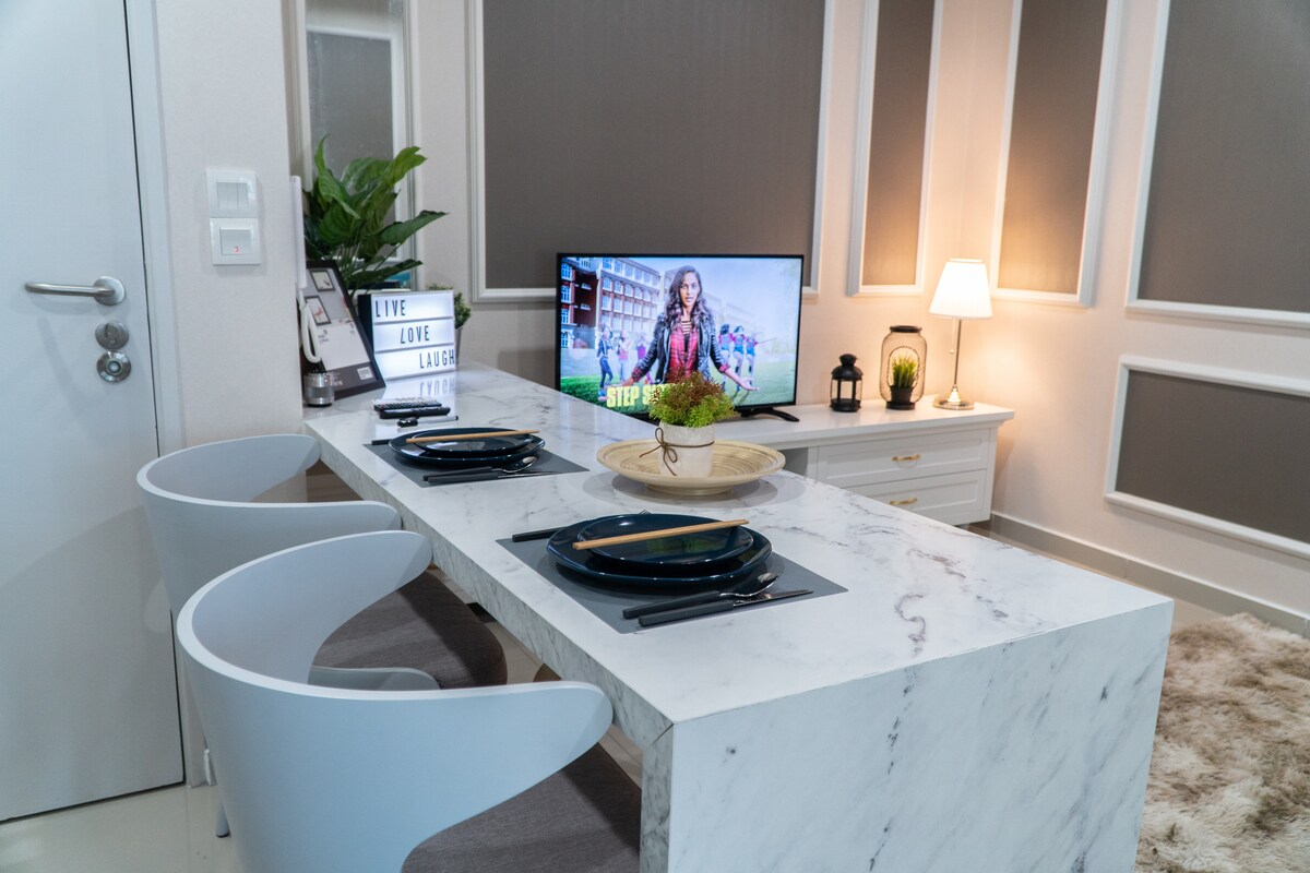 A modern dining area features a white marble table with two sleek chairs. A television displays vibrant colors, while a soft lamp and decorative items create a cozy ambiance. A potted plant adds a touch of greenery to the well-lit space.