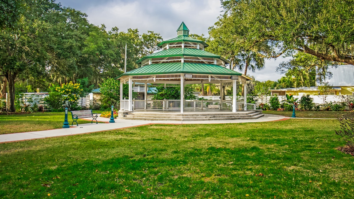 A charming gazebo features an intricate design with a green roof, surrounded by well-maintained grass and trees. A paved walkway leads to the structure, and nearby benches provide places to sit and enjoy the outdoor scenery. Lush landscaping enhances the peaceful setting.