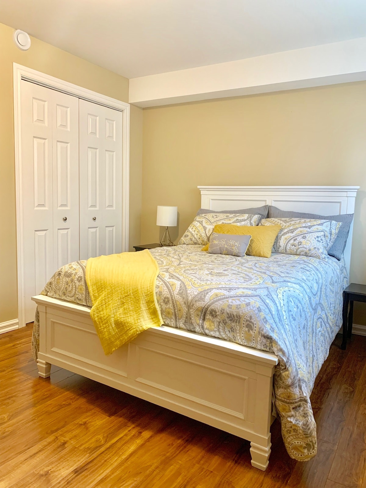A well-lit bedroom features a spacious bed adorned with an intricate patterned comforter and matching pillows. A nightstand holds a lamp beside the bed, while double doors lead to a closet, providing ample storage space. Warm wooden flooring complements the neutral-toned walls.