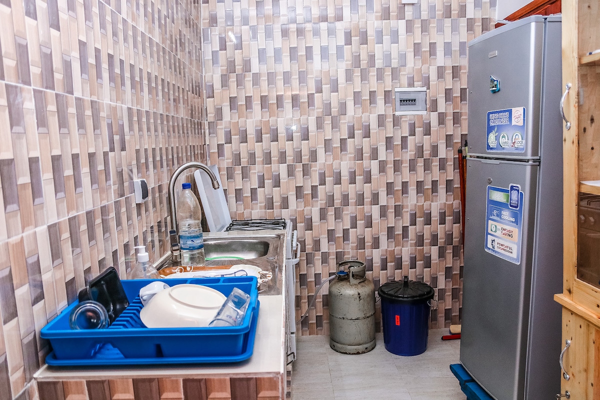 A compact kitchen area features a stainless steel refrigerator and a gas cylinder, with a sink and countertop space. Dishware is arranged in a blue tray, while containers are stored nearby. The wall showcases a modern tile pattern in neutral tones.