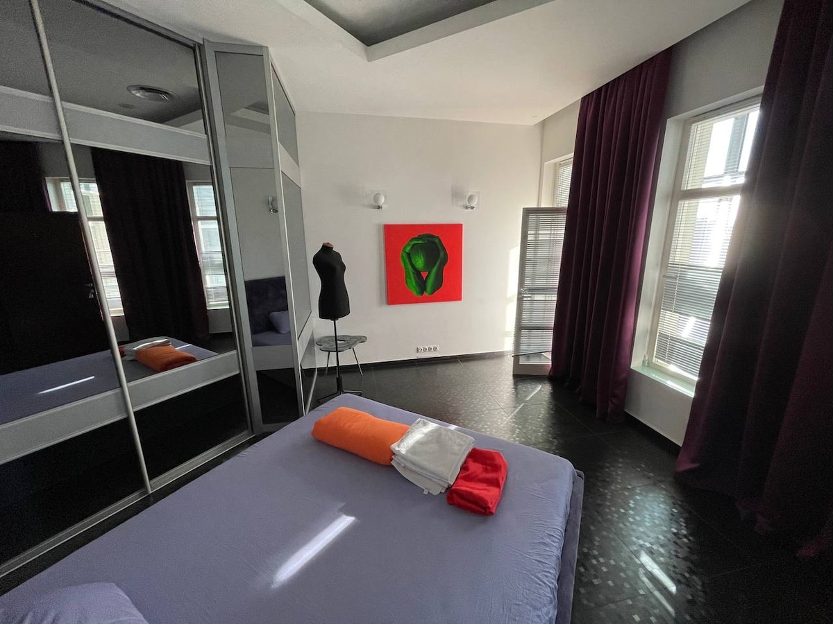 A bright bedroom features large windows with natural light illuminating the space. A spacious bed is adorned with neatly arranged orange and red blankets. A colorful artwork adorns the wall, and a standing mirror and a clothing mannequin add functional elements to the room.
