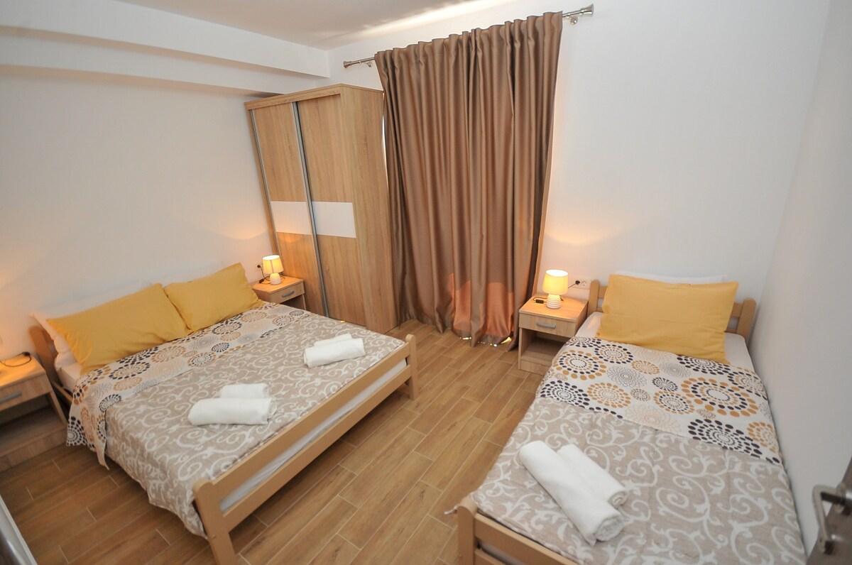 A well-arranged sleeping area features two beds with light yellow bedding and patterned throws. Soft lighting is provided by bedside lamps on wooden nightstands. Light brown tiled flooring complements the warm tones in the space, while curtains in a muted shade frame a window.