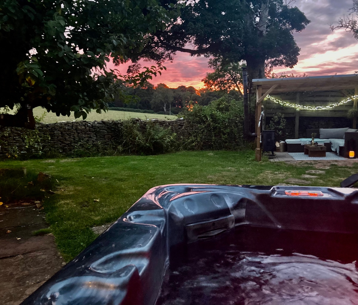A hot tub is positioned in a landscaped garden, surrounded by greenery. In the background, a sunset casts warm hues across an open field, while a cozy seating area with ambient lighting is visible beneath a covered patio.