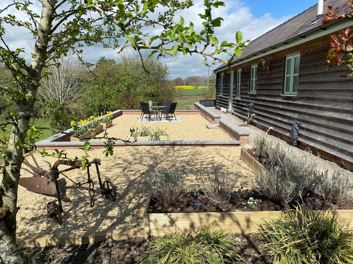 A gravel patio area is seen outside the bungalow, featuring a set of four black chairs and a round table. Surrounding garden beds, planted with flowers and shrubs, add greenery, while the distant hills are visible under a partly cloudy sky.