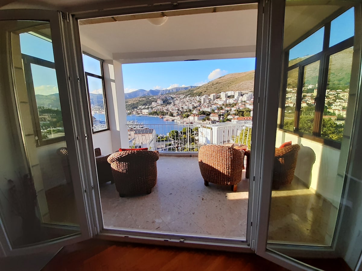 An inviting balcony creates a relaxing space with woven seating, offering panoramic views of the harbor and surrounding hills. Large glass doors open to the outside, allowing natural light to illuminate the area and connect the indoors with the breathtaking scenery.