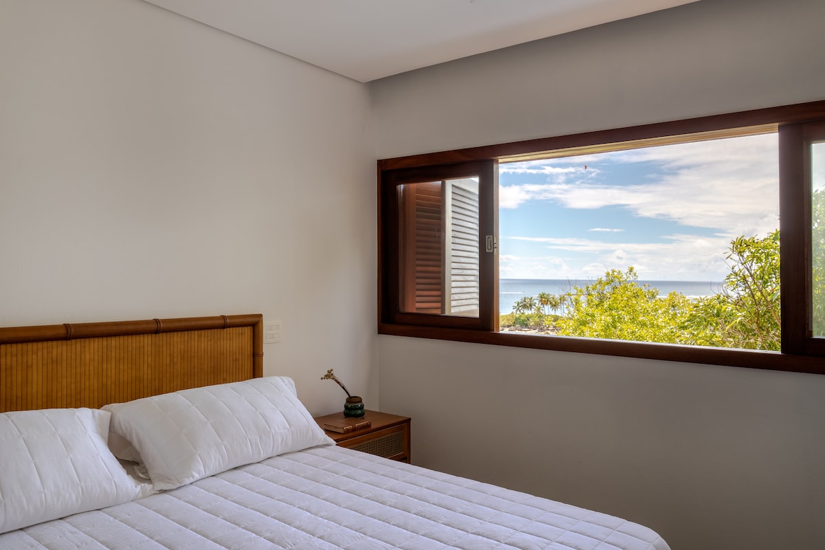 A spacious bedroom features a comfortable Queen bed with crisp white linens. A large window offers a view of the greenery and the ocean beyond, while natural light brightens the serene space. A nightstand with a small lamp is positioned beside the bed.