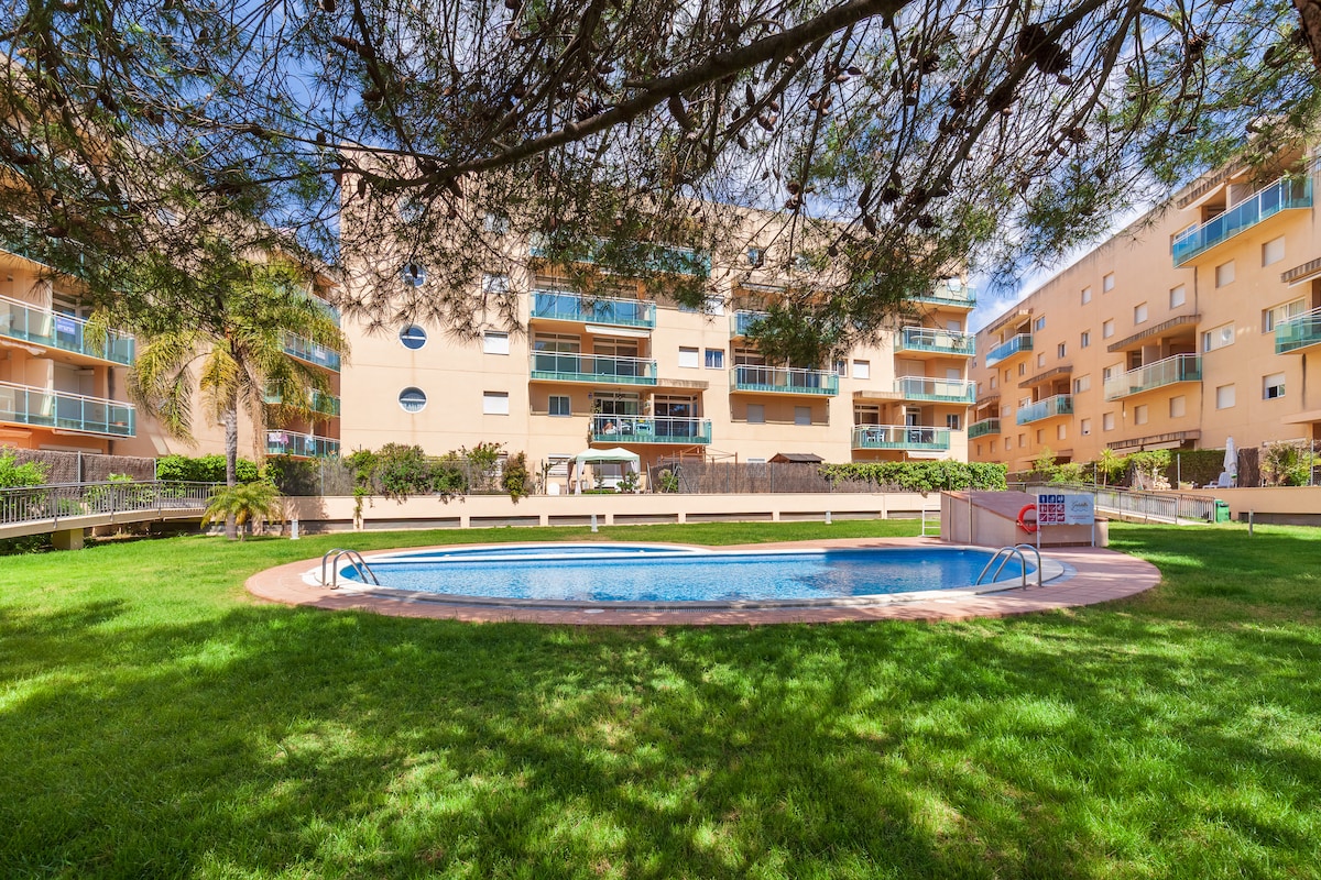 A community pool is situated within a grassy area surrounded by residential buildings. Sun loungers are positioned near the pool, which is gently shaded by trees. Clear blue water can be seen invitingly glistening under the sunlight.