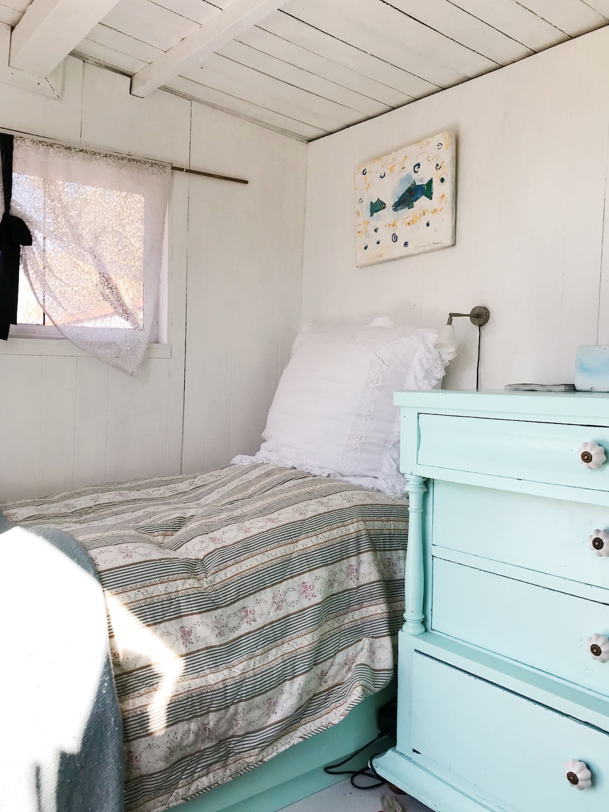 A cozy sleeping area features a single bed with a textured quilt and a white pillow. A turquoise nightstand with drawers is positioned beside the bed. Natural light streams in through a window adorned with a light curtain, illuminating the simple decor.