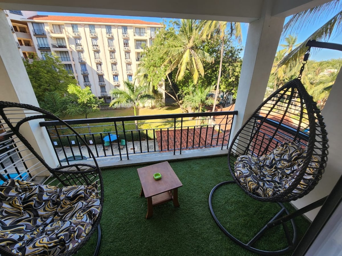 A balcony space is equipped with two hanging chairs, each featuring patterned cushions. A small wooden table sits between the chairs. Lush greenery and palm trees are visible in the background, along with a glimpse of the building's exterior across the way.