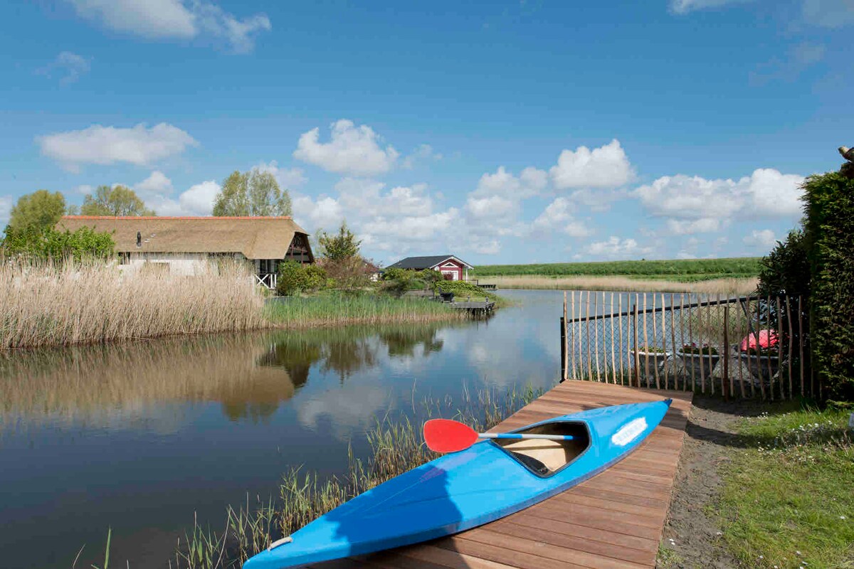 A brightly colored blue kayak rests on a wooden dock, positioned alongside a serene waterway. Lush greenery and tall reeds frame the scene, while fluffy clouds drift across a clear blue sky, creating a peaceful atmosphere at the water's edge.