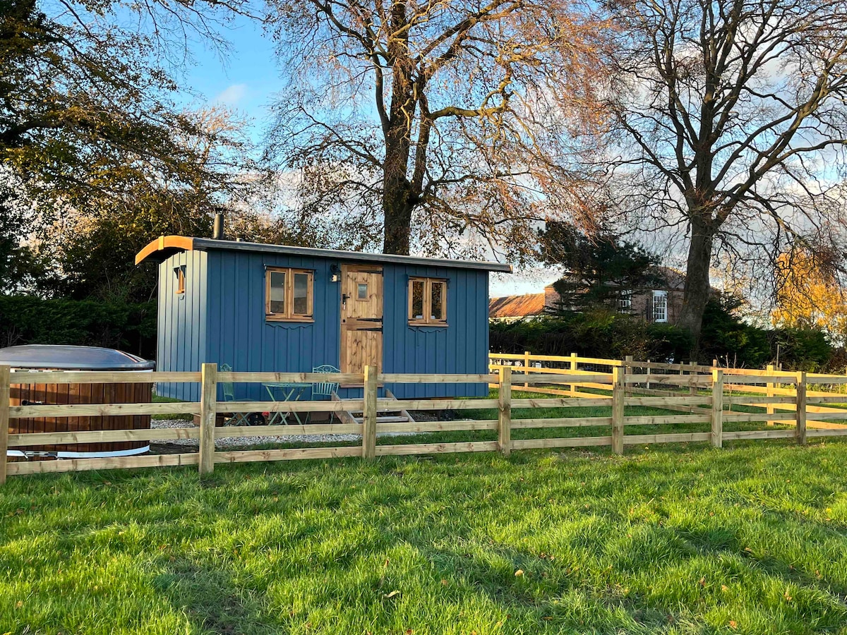 A charming blue shepherd's hut is set within a fenced grassy area, surrounded by trees. A wood-fired hot tub is positioned nearby, inviting outdoor relaxation. The entrance features wooden doors and two small windows, allowing natural light to enhance the hut's cozy appearance.