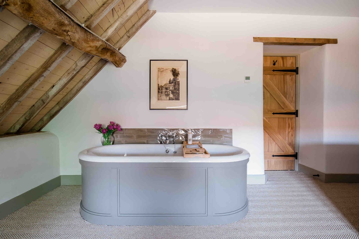 A freestanding tub is positioned under a sloped wooden ceiling, complemented by exposed beams. A bouquet of flowers decorates the tub's edge, and a piece of framed artwork is displayed above it. Warm tones from the wooden door enhance the serene atmosphere.