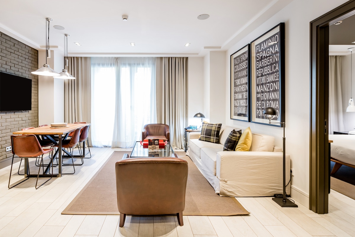 A spacious living area features large windows allowing natural light to fill the room. A comfortable sofa and leather chairs surround a glass coffee table. A dining table with several chairs is positioned near the window, with modern artwork adorning the walls.