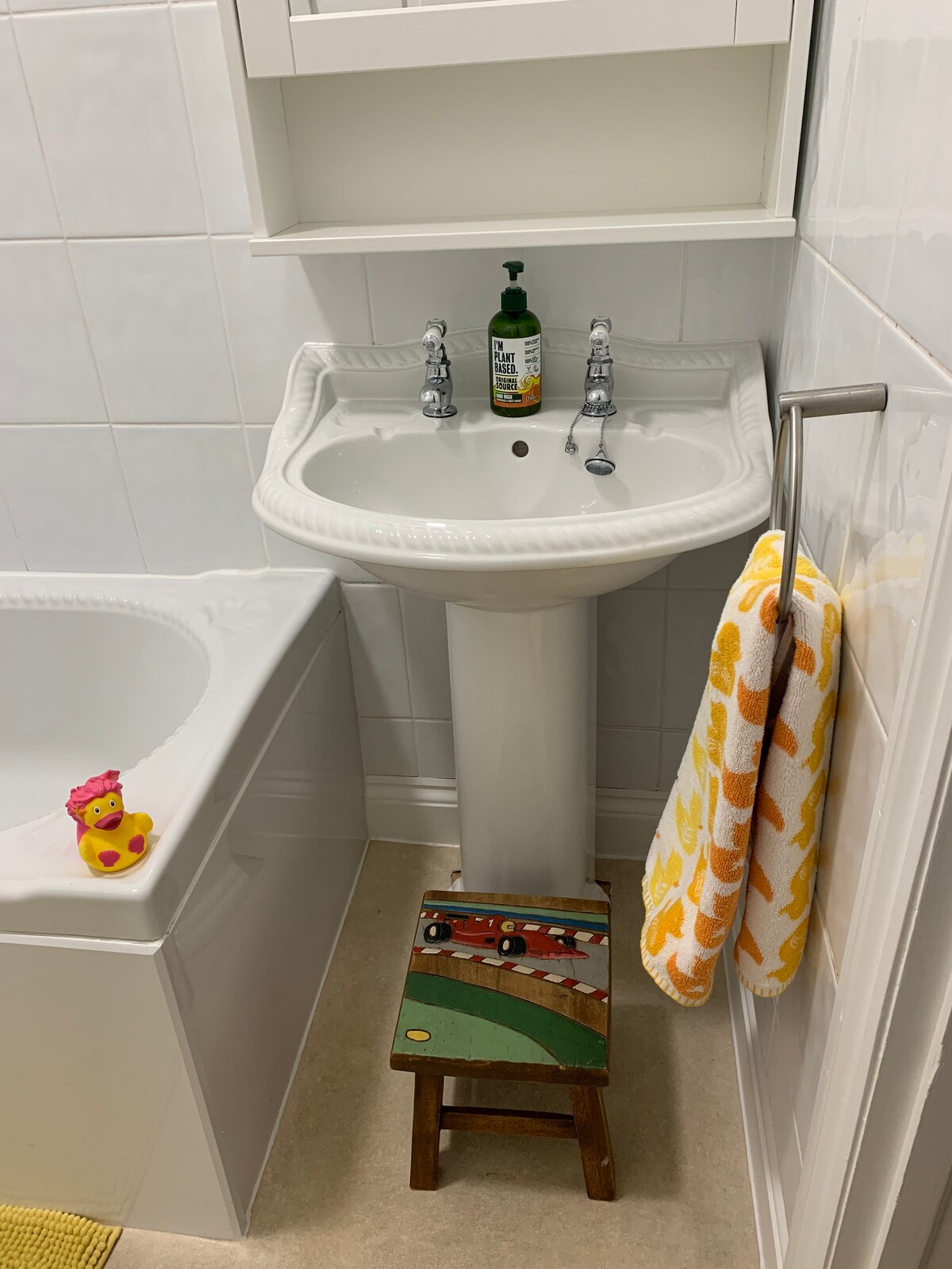 A compact bathroom is displayed, featuring a porcelain sink with traditional taps. A small wooden stool is positioned beneath the sink. A yellow and white patterned towel hangs from a chrome rail, while a rubber duck rests beside the sink.