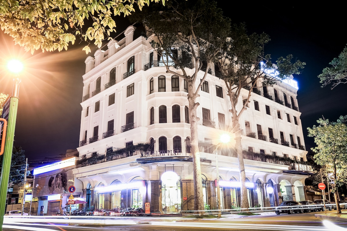 The elegant five-story building is illuminated at night, showcasing its architectural details. Large windows and balconies are positioned along each floor, while tall trees line the street, providing a natural setting. Soft lighting enhances the welcoming ambiance of the entrance.