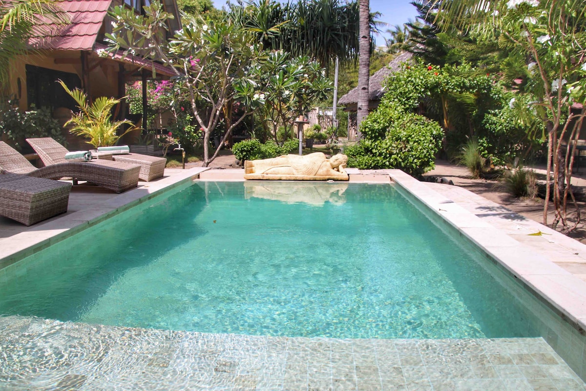 A serene pool area is framed by lush tropical gardens. Sunbeds with soft cushions are positioned nearby, offering a comfortable place to relax. A tranquil statue is visible at the pool's edge, enhancing the peaceful atmosphere of the setting.