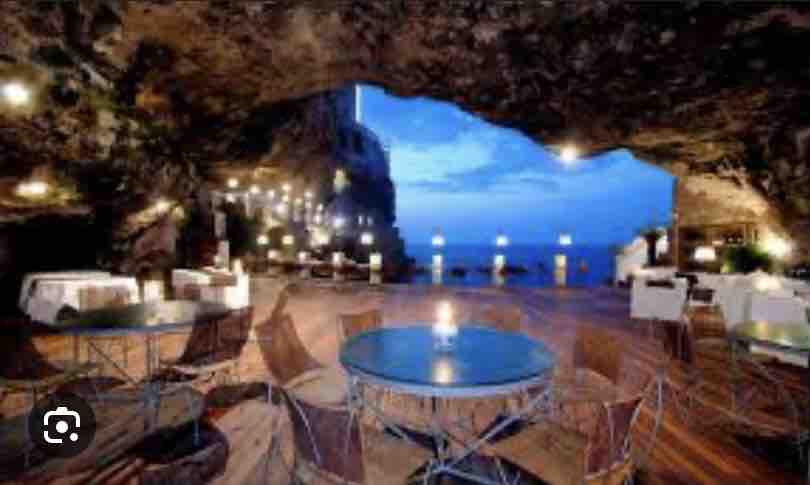 A cave dining area is illuminated by soft lights hanging from the rocky ceiling. Tables are set with candles, surrounded by elegant chairs. A view of the sea is visible through the opening, with twilight sky enhancing the ambiance.