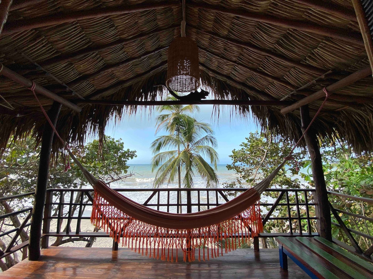 A shaded sitting area features a hammock suspended between wooden posts, overlooking lush greenery and the ocean. The thatched roof adds a natural touch, while a light fixture hangs above, enhancing the tranquil atmosphere of this beachfront escape.