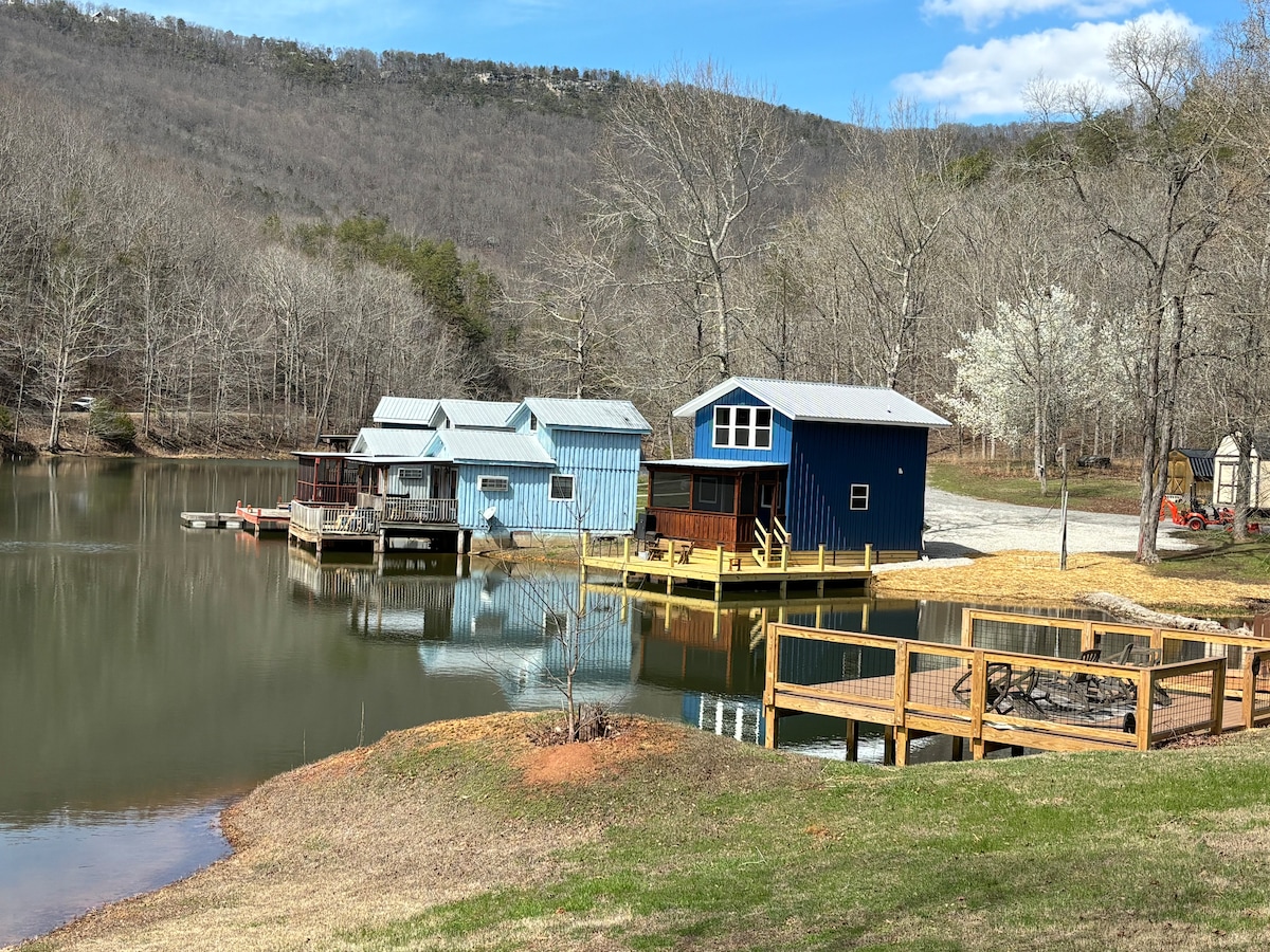Three waterfront cabins are situated by a serene lake, reflecting the surrounding landscape. A wooden deck with seating provides a space for relaxation, while the backdrop features gently sloping hills and bare trees. Sunlight illuminates the scene against a clear blue sky.
