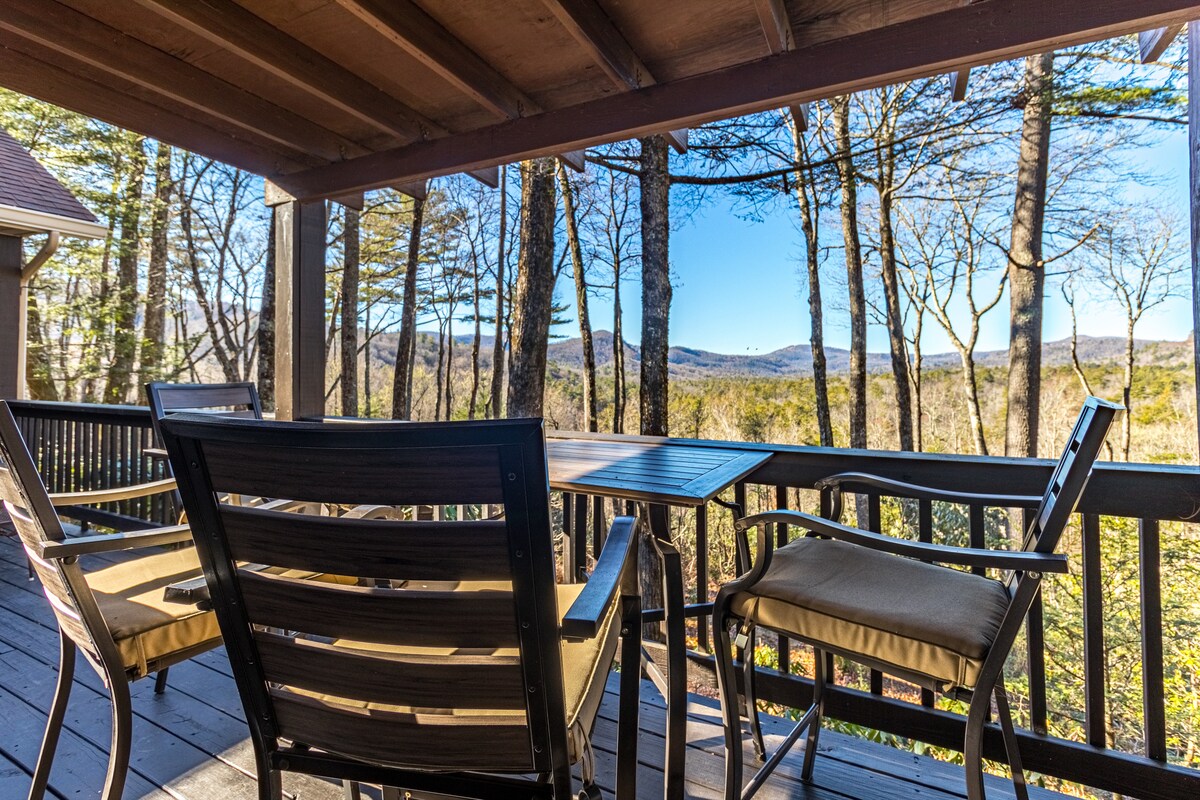 A spacious deck is furnished with four comfortable chairs surrounding a table, offering views of the wooded landscape and distant mountains. Natural light fills the area, creating an inviting outdoors space designed for relaxation.