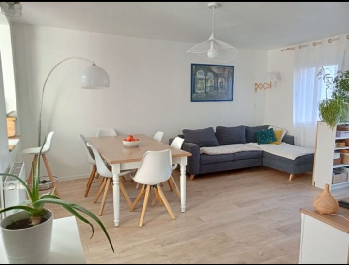 A bright and airy living space features a light wooden floor, a large dining table surrounded by four chairs, and a comfortable sectional sofa adorned with colorful cushions. A piece of artwork hangs on the wall, while natural light streams through sheer window coverings.