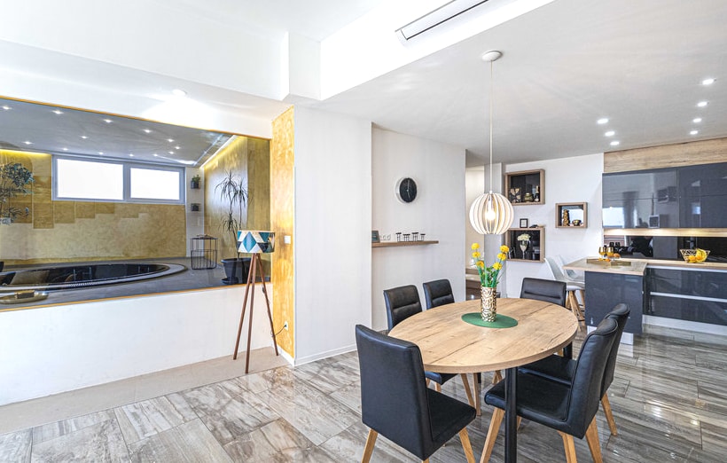 A modern dining area features a round wooden table surrounded by black chairs. A stylish pendant light hangs above. In the background, an indoor hot tub is visible, flanked by light-colored walls that create an open and spacious feel.