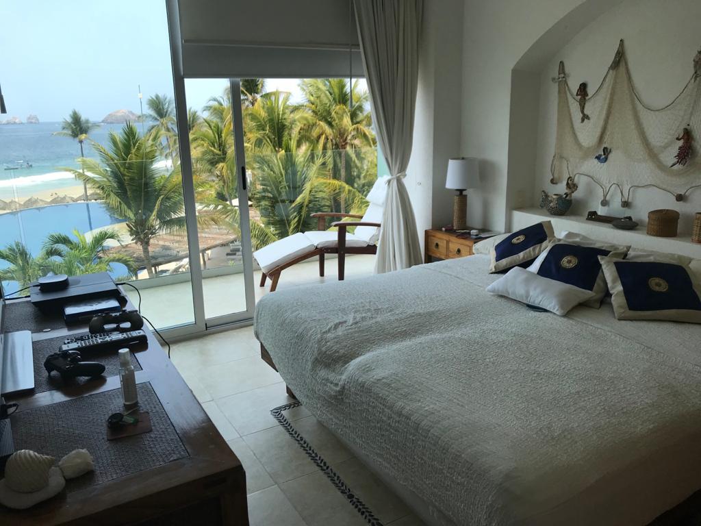 A spacious bedroom is featured, showcasing a large bed adorned with decorative pillows. Natural light floods the room through a glass door leading to a view of palm trees and the ocean. A cozy seating area is positioned beside the window.